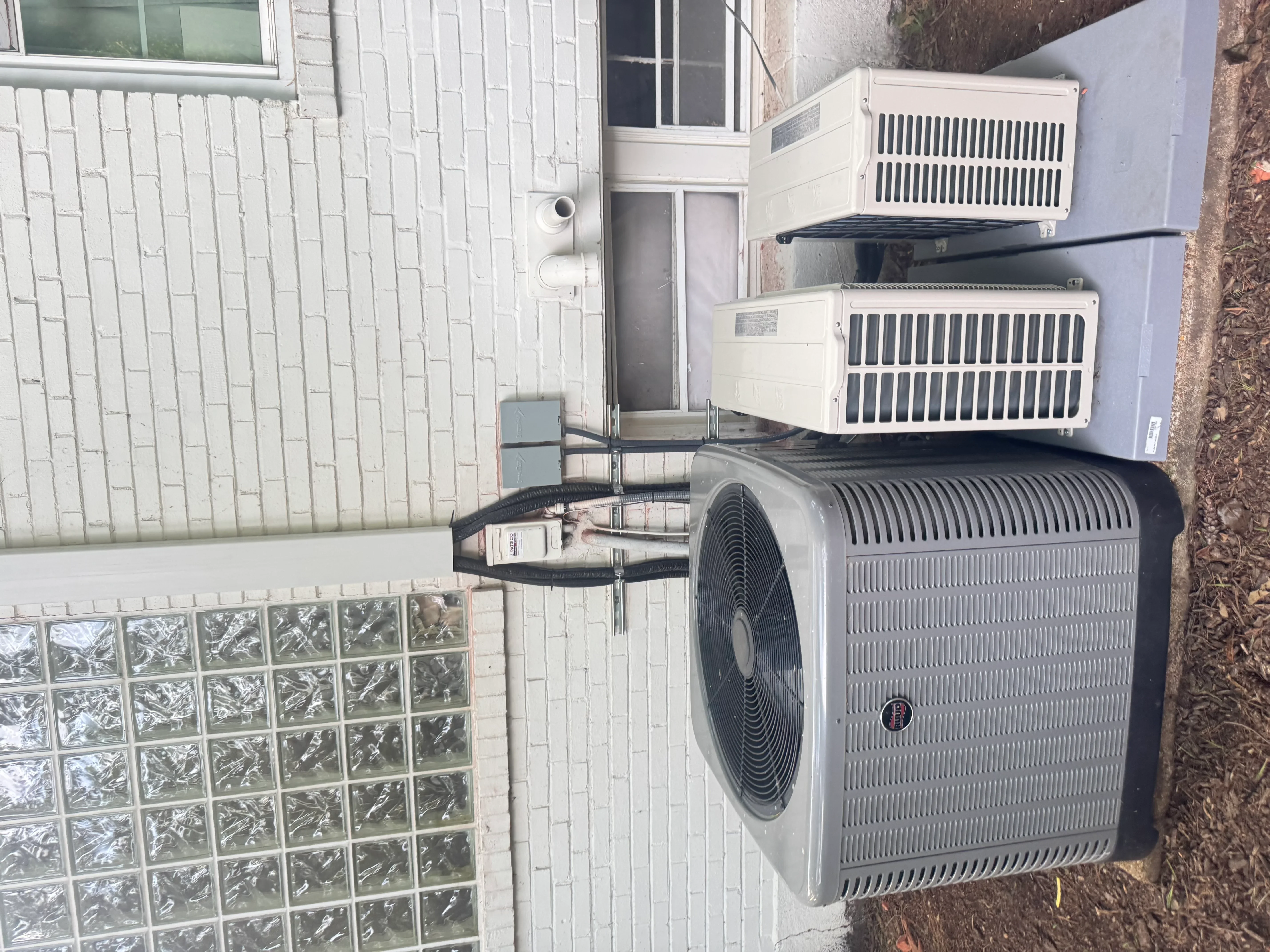 Outdoor HVAC condenser unit with refrigerant lines and three interior air handler units mounted on white brick wall