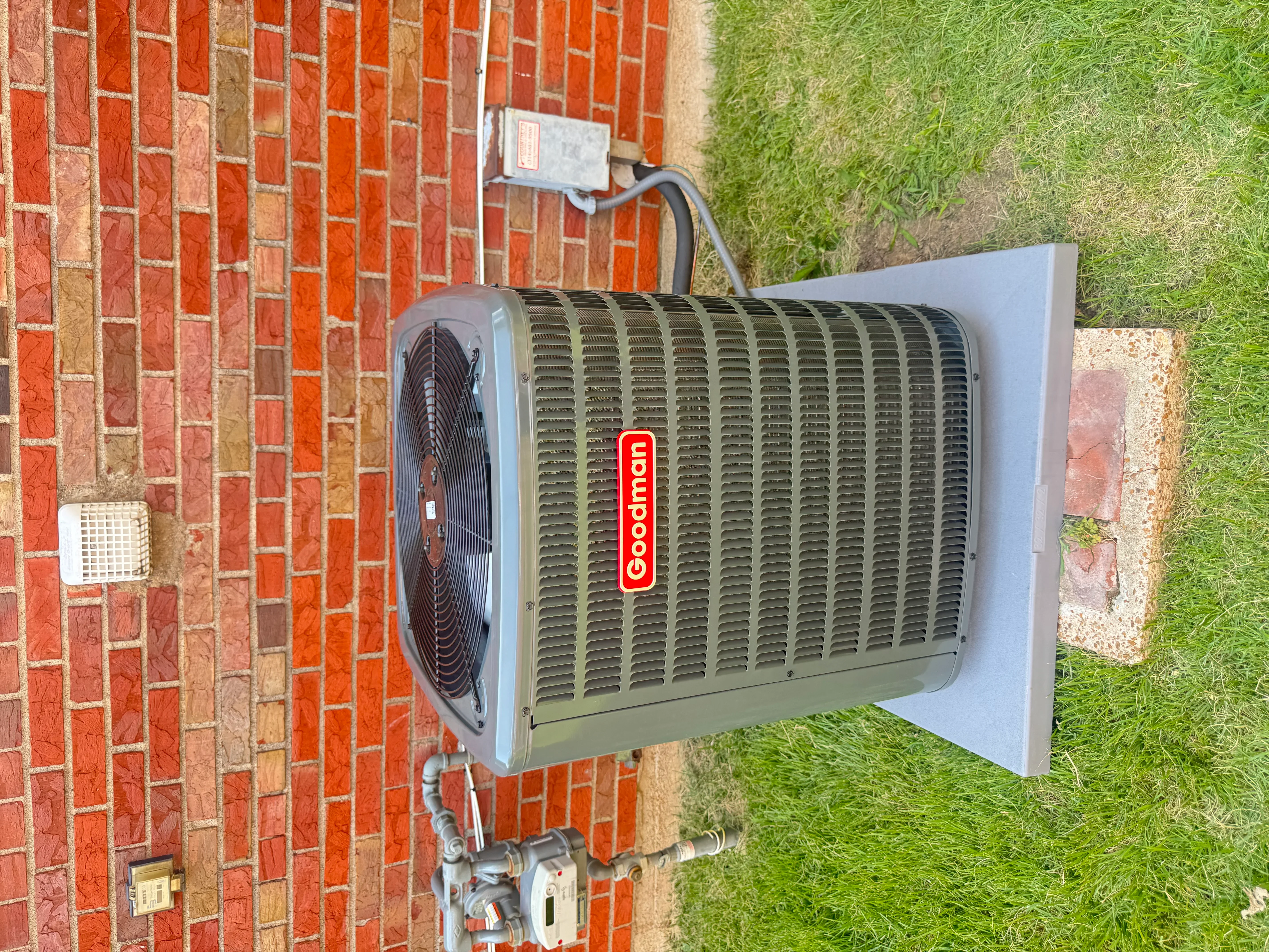 Goodman AC condenser unit mounted on red brick wall with electrical disconnect and refrigerant lines