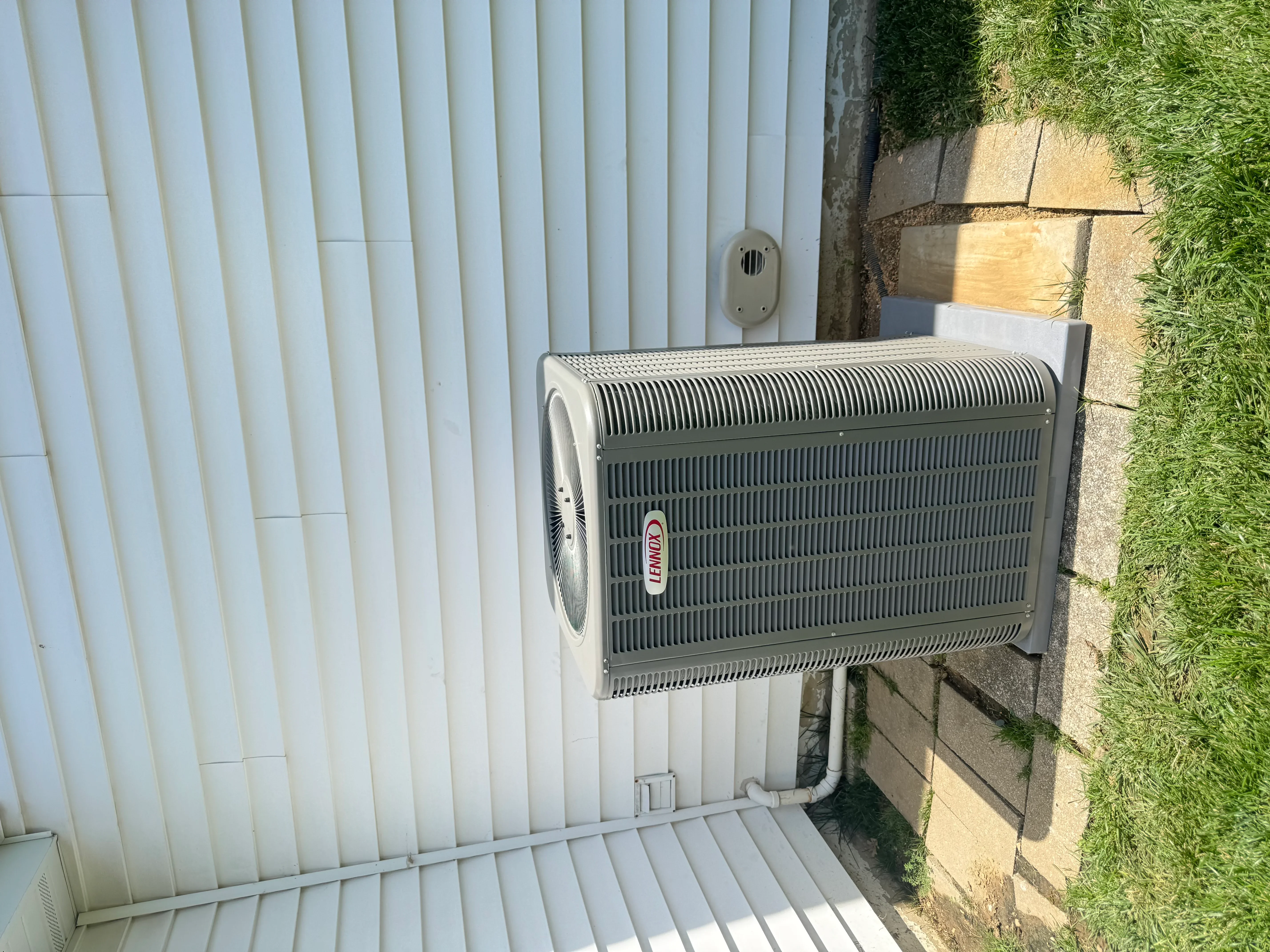 Lennox AC condenser unit mounted on white metal siding with refrigerant lines and electrical connections